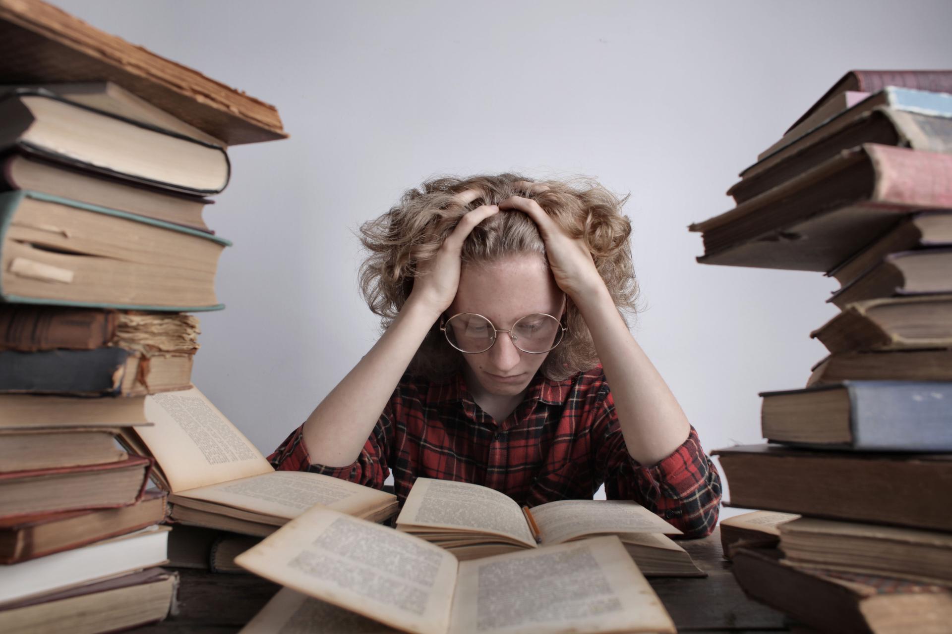 Teenage boy with glasses stressing over homework surrounded by stacks of books indoors.